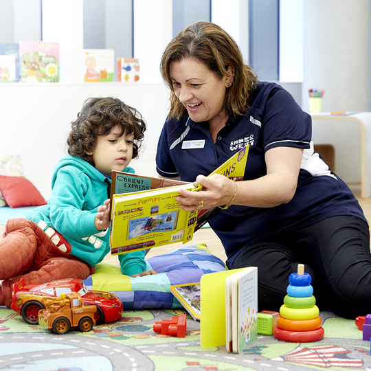 Woman sitting on the floor with a child reading a book together with toys scattered around